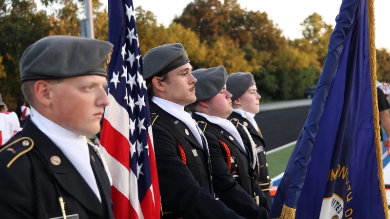 ROTC cadets in uniform stand at attention during a flag ceremony, holding the American and military service flags