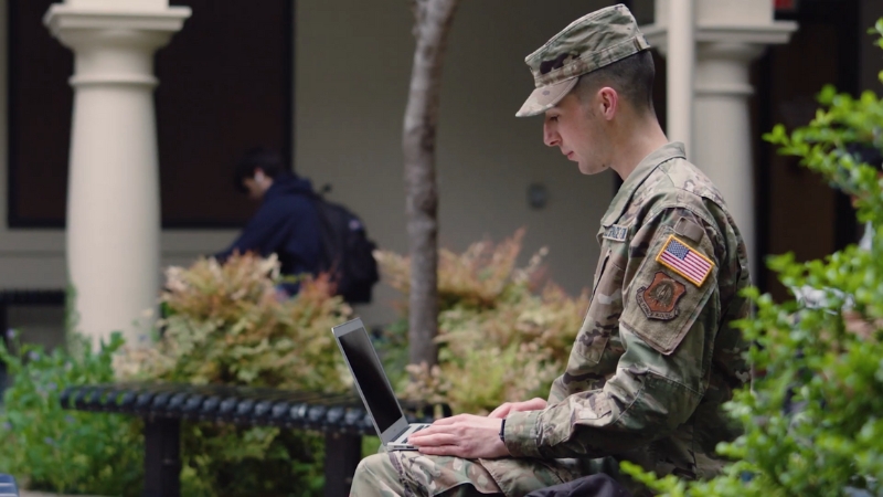 An ROTC cadet in uniform works on a laptop while seated outside on campus