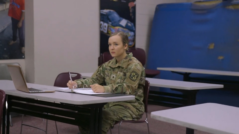 A female ROTC cadet in uniform takes notes during class, with a laptop and binder open on the desk