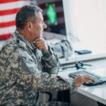 A US Army soldier in uniform works on a computer at a desk with an American flag in the background