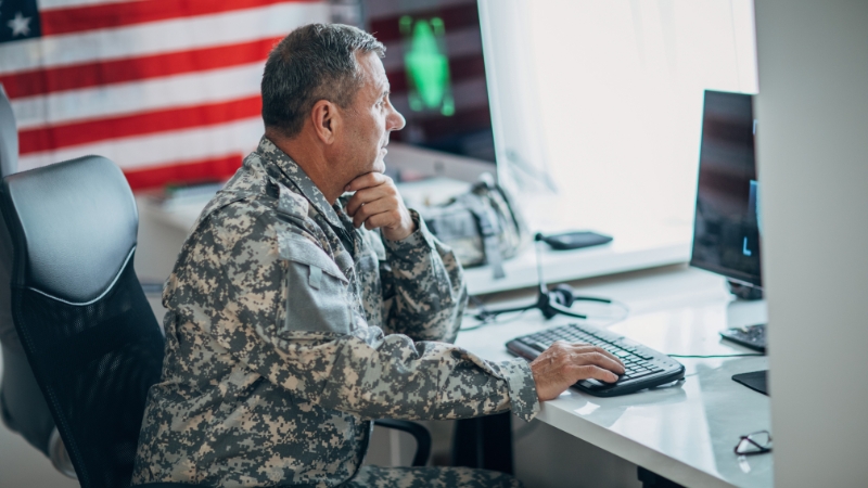 A US Army soldier in uniform works on a computer at a desk with an American flag in the background