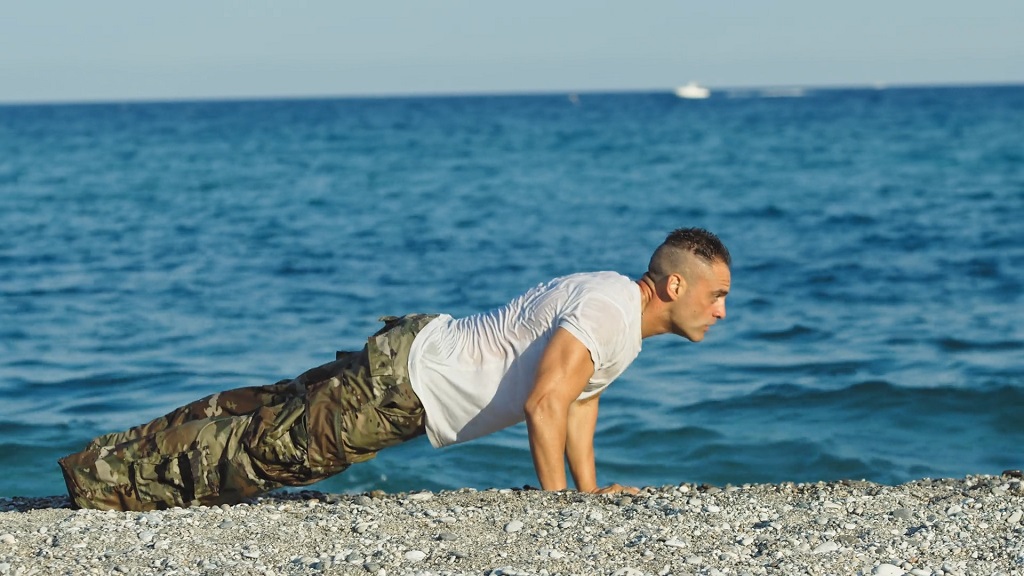 Man in camouflage pants and a white t-shirt doing push-ups on a beach with the sea in the background, symbolizing the influence of military-style fitness in popular culture.