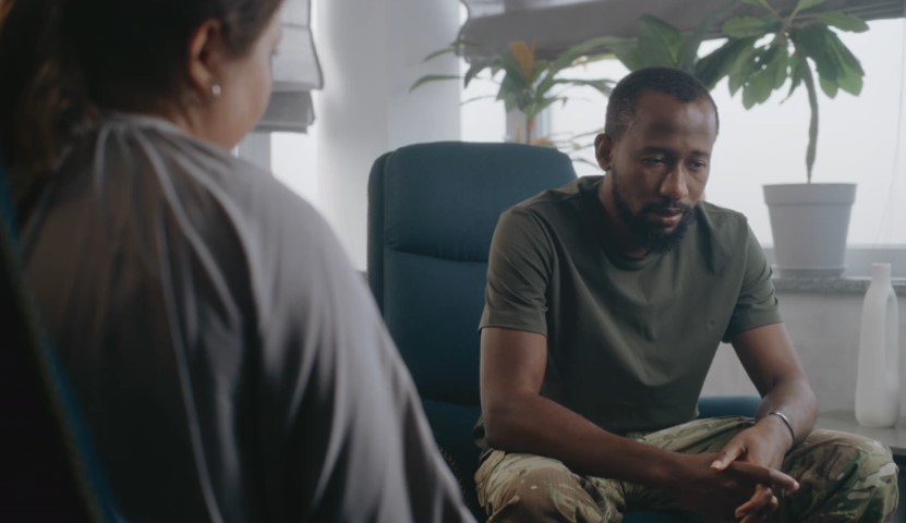 A man in military-style pants and an olive green t-shirt sits with his hands clasped, looking down as if in deep thought during a session with a therapist