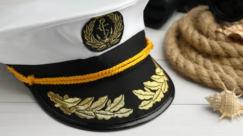 Navy officer cap with gold embroidery placed on a table beside rope and seashells