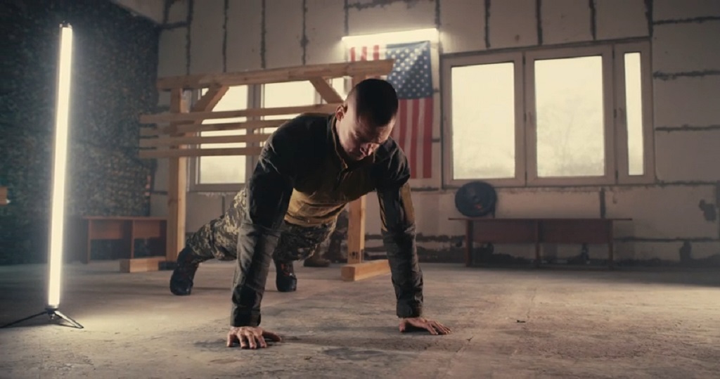 Soldier in military uniform doing push-ups indoors with an American flag in the background, symbolizing strength and resilience