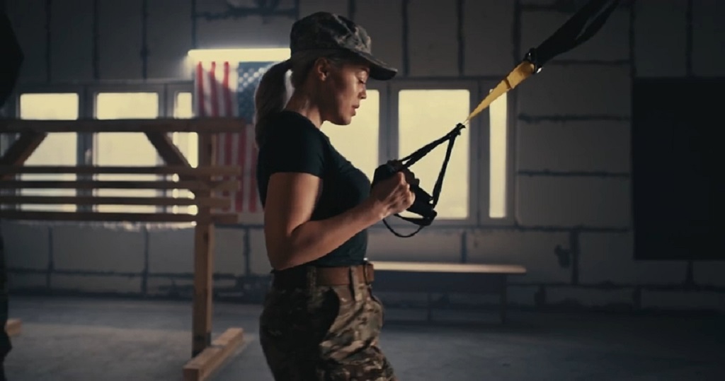 Woman in military-style training gear using suspension straps indoors, with the U.S. flag in the background, symbolizing civilian adoption of military fitness methods