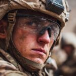 Close-up of a soldier wearing tactical gear, goggles, and a helmet, with dust on his face and other soldiers blurred in the background